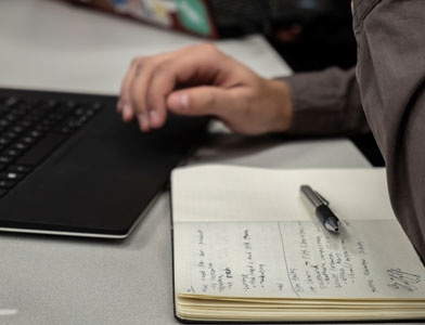 A person's hand is shown at a laptop with a journal and pen in the foreground